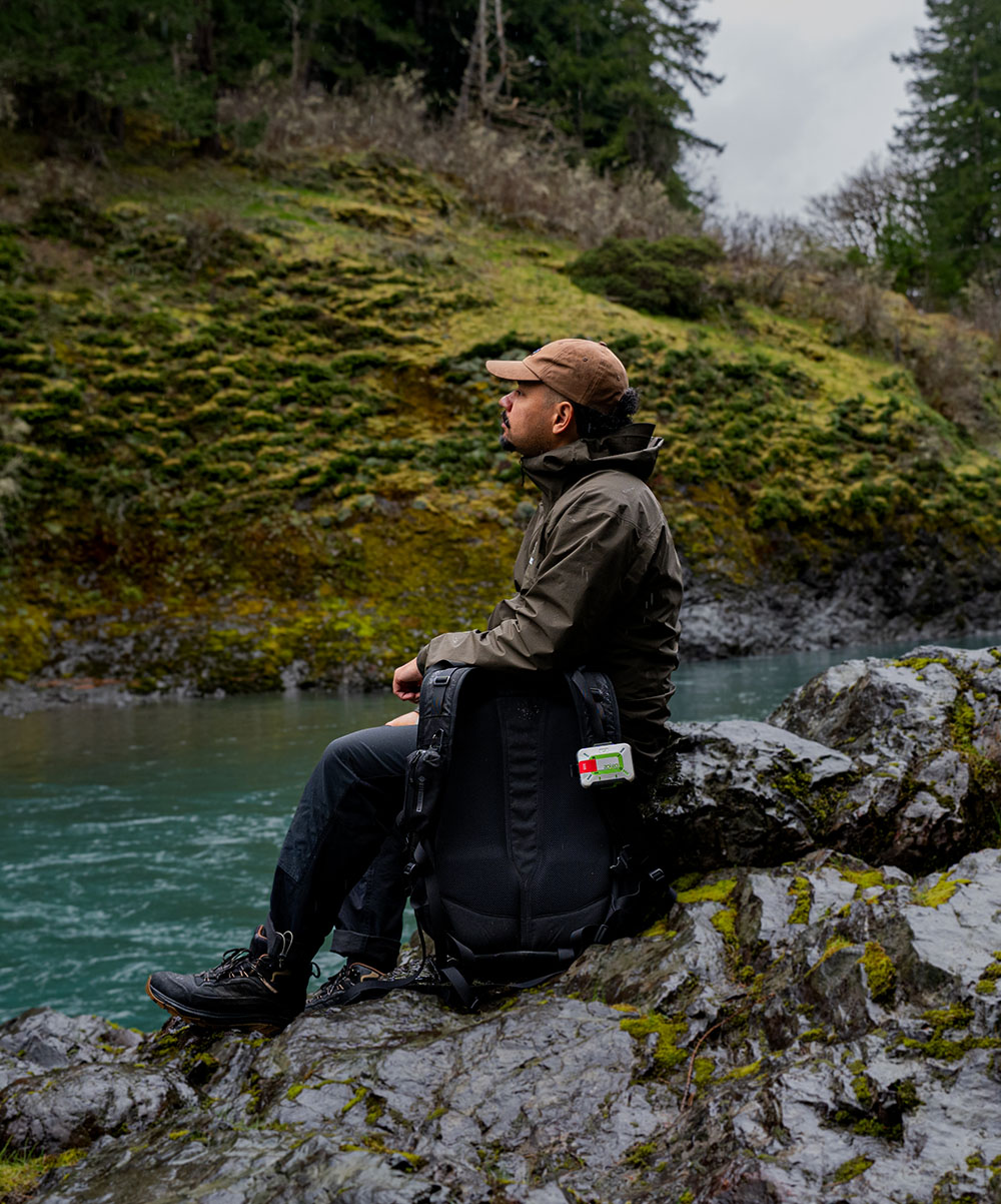 A hiker resting beside a river with a ZOLEO communicator clipped to his backpack